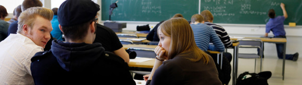 Students sitting in a classroom, someone half-kneeling in the background writing on the blackboard